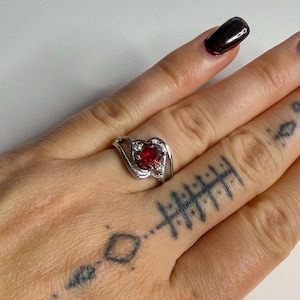 Peut inclure: Une bague en argent avec une pierre rouge en forme de cœur au centre. La bague présente un motif tourbillonnant avec de petites pierres d'accent claires. La bague est sur le doigt d'une personne.