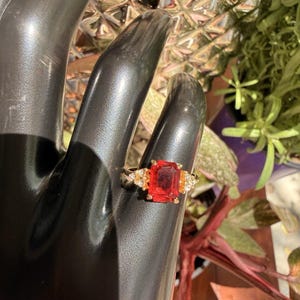 Könnte beinhalten: Ein goldener Ring mit einem großen, rechteckigen, roten Edelstein im Mittelpunkt. Der Ring wird von kleineren, runden, gelben Edelsteinen auf jeder Seite flankiert. Der Ring wird auf einer schwarzen Hand präsentiert.