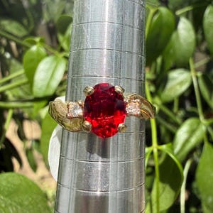 May include: A gold ring featuring a large, oval-cut, deep red gemstone at the center, flanked by two smaller, round, clear stones. The band has a textured, brushed finish and is displayed on a silver-toned, cylindrical ring sizing mandrel.