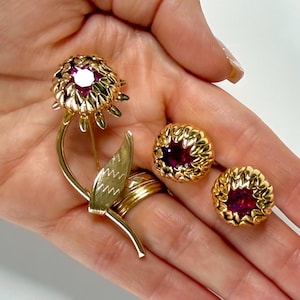 May include: A gold-toned floral brooch and matching earrings set. The brooch is a flower with a central red gemstone and detailed petals. The earrings are round with a similar design and red gemstone centers. Jewelry.