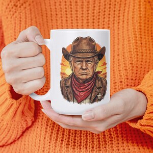 Puede incluir: Taza de cerámica blanca con una ilustración de vaquero. El diseño presenta a un hombre con sombrero de vaquero marrón, pañuelo rojo y chaqueta de cuero marrón, sobre un fondo de puesta de sol.