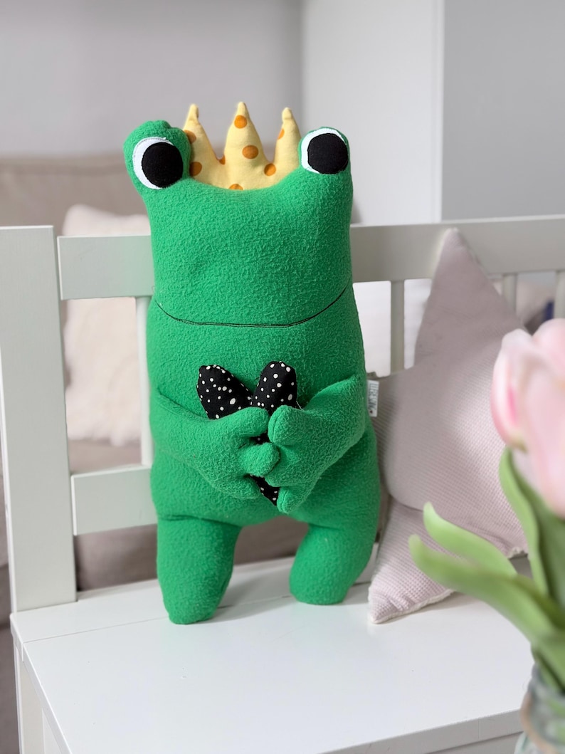 May include: A green plush frog toy wearing a yellow crown with orange spots. The frog has large black eyes and holds a black and white polka dot heart. The toy is on a white surface.