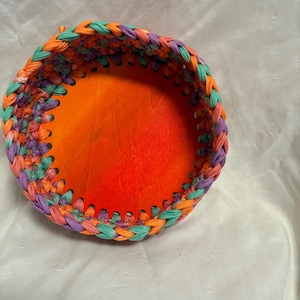 Puede incluir: Una cesta redonda, hecha a mano, con una combinación de colores vibrantes. El borde de la cesta está trenzado con tiras de tela en tonos naranja, morado y turquesa. El interior es de color naranja sólido, creando un contraste llamativo. Aproximadamente 15 cm de diámetro.