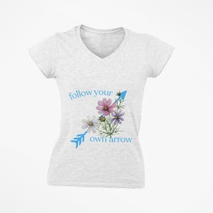 May include: A light gray V-neck t-shirt with the text "follow your own arrow" in light blue. The shirt features a floral design with pink, purple, and white flowers and a blue arrow.