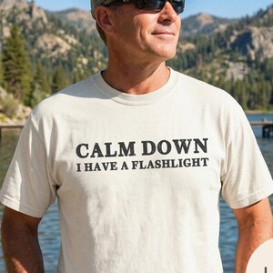 Puede incluir: Camiseta color marfil con el texto negro "CALM DOWN I HAVE A FLASHLIGHT". La persona lleva gafas de sol y una gorra verde. El fondo muestra un lago y montañas.