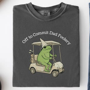 Peut inclure: T-shirt de couleur poivre avec une grenouille de dessin animé conduisant un chariot de golf, portant des lunettes de soleil et un chapeau. Le texte "Off to Commit Dad Foolery" est au-dessus de la grenouille. Le t-shirt est présenté avec un jean bleu et une serviette à motifs.