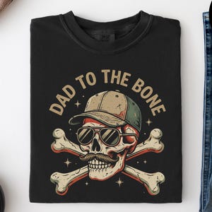 Peut inclure: T-shirt noir avec un motif de crâne portant des lunettes de soleil et une casquette de baseball, avec des os croisés et le texte "DAD TO THE BONE". Le design est de style vintage. Le t-shirt est plié sur une surface blanche.
