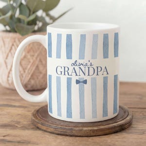 Peut inclure: Mug en céramique blanche avec des rayures verticales bleues et le texte "olivia's GRANDPA" en bleu marine. Un petit nœud papillon bleu est en dessous du texte. Le mug est posé sur un dessous de verre en bois.