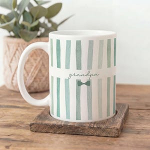 Peut inclure: Mug en céramique blanche avec des rayures verticales turquoise et le mot "grandpa" en écriture cursive. Un motif de nœud papillon turquoise est en dessous. Le mug est posé sur un petit dessous de verre carré en bois.