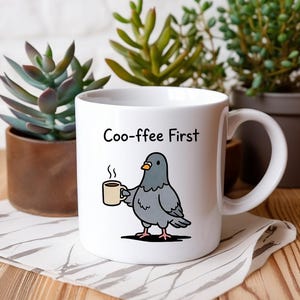 Op de afbeelding: Witte keramische mok met de tekst "Coo-ffee First" en een cartoon duif met een koffiekopje. De mok staat op een patroon doek, met potplanten op de achtergrond.