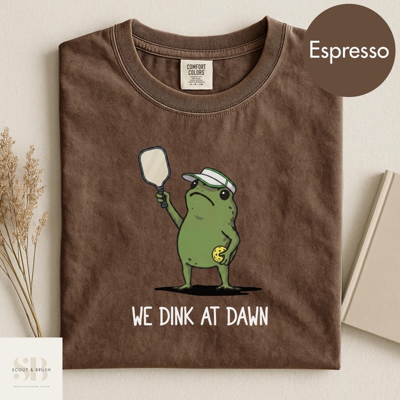 K&ouml;nnte beinhalten: Espressofarbenes T-Shirt mit einem Cartoon-Frosch, der Pickleball spielt. Der Frosch tr&auml;gt ein Visier und h&auml;lt einen Schl&auml;ger, mit einem Pickleball in der anderen Hand. Der Text "WE DINK AT DAWN" ist darunter gedruckt.