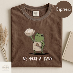 May include: Espresso-colored t-shirt featuring a cartoon frog chef holding bread in a basket and a wooden spoon. The text "WE PROOF AT DAWN" is printed below the frog. The shirt is made of Comfort Colors material.