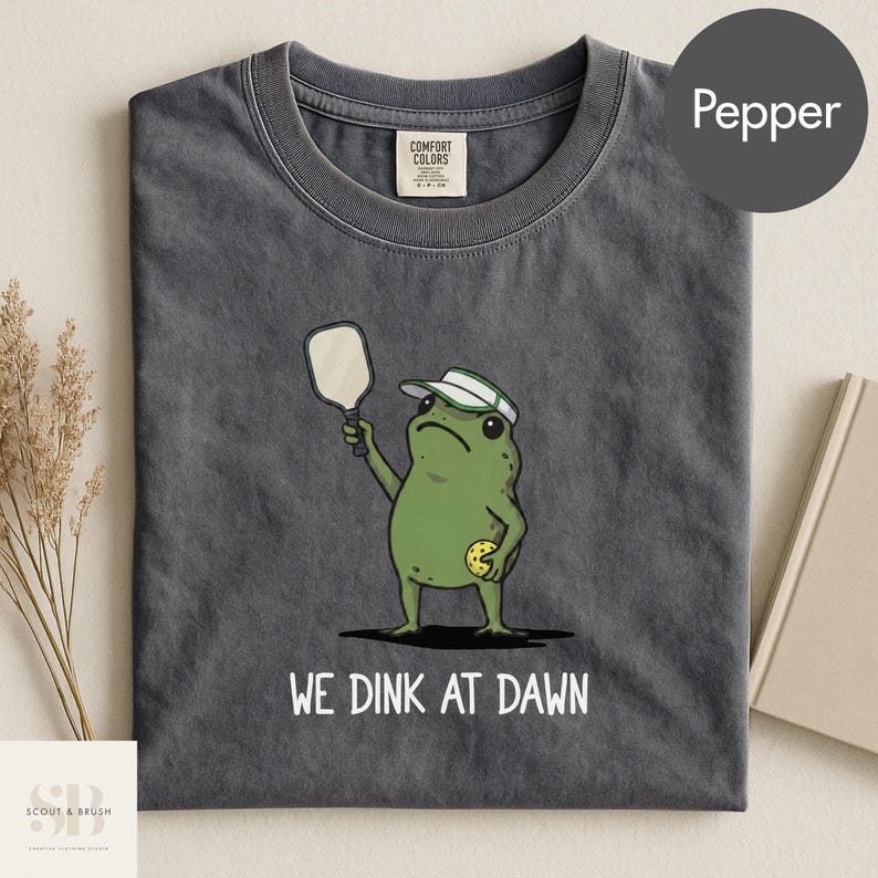 K&ouml;nnte beinhalten: Pfefferfarbenes T-Shirt mit einem gr&uuml;nen Frosch, der einen Schirmm&uuml;tze tr&auml;gt und einen Pickleball-Schl&auml;ger und -Ball h&auml;lt. Der Text "WE DINK AT DAWN" ist darunter gedruckt. Das Shirt hat einen Rundhalsausschnitt und kurze &Auml;rmel.