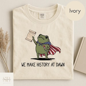 Könnte beinhalten: Elfenbeinfarbenes T-Shirt mit einer Cartoon-Froschfigur, die einen Umhang trägt und eine Schriftrolle und ein Feuerwerk hält. Der Frosch ist grün mit einem blau-weißen Schal. Der Text "WE MAKE HISTORY AT DAWN" ist darunter gedruckt.