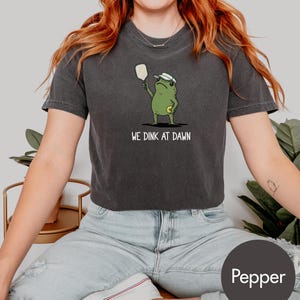 K&ouml;nnte beinhalten: Dunkelgraues T-Shirt mit einer Cartoon-Frosch, der Pickleball spielt. Der Frosch tr&auml;gt ein Visier und h&auml;lt einen Schl&auml;ger, darunter der Text "WE DINK AT DAWN". Das Shirt hat die Farbe Pfeffer.