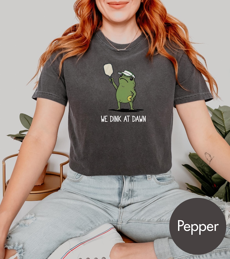 K&ouml;nnte beinhalten: Dunkelgraues T-Shirt mit einer Cartoon-Frosch, der Pickleball spielt. Der Frosch tr&auml;gt ein Visier und h&auml;lt einen Schl&auml;ger, darunter der Text "WE DINK AT DAWN". Das Shirt hat die Farbe Pfeffer.