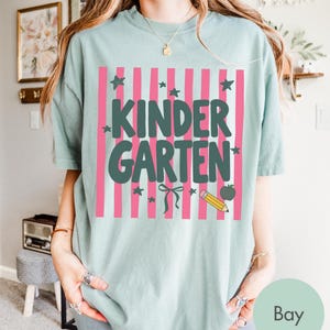 May include: A sage green short-sleeve t-shirt featuring a graphic print of vertical pink stripes. The text "KINDERGARTEN" is printed in dark green block letters, accompanied by small dark green stars, a bow, a yellow pencil, and an apple.