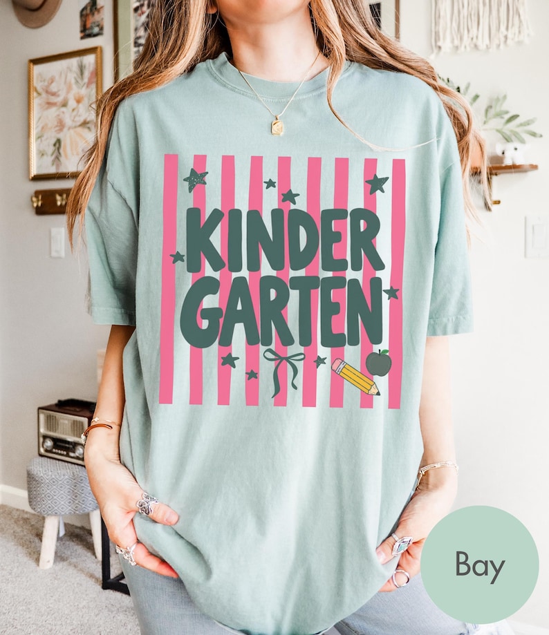 May include: A sage green short-sleeve t-shirt featuring a graphic print of vertical pink stripes. The text "KINDERGARTEN" is printed in dark green block letters, accompanied by small dark green stars, a bow, a yellow pencil, and an apple.