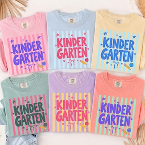 May include: Six crew neck t-shirts in pink, light blue, yellow, sage green, lavender, and coral. Each shirt features a vertical striped background with the text KINDER GARTEN in bold letters, surrounded by small star, pencil, apple, and ribbon graphics.