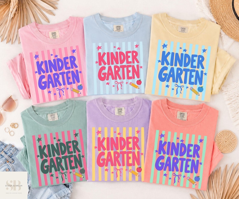 May include: Six crew neck t-shirts in pink, light blue, yellow, sage green, lavender, and coral. Each shirt features a vertical striped background with the text KINDER GARTEN in bold letters, surrounded by small star, pencil, apple, and ribbon graphics.