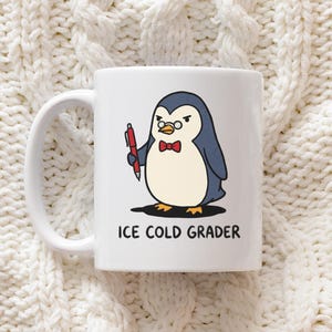 May include: White ceramic mug featuring a cartoon penguin with glasses, a bow tie, and a pen. The penguin is labeled "ICE COLD GRADER" in black text. The mug has a white handle and is set against a white knitted background.