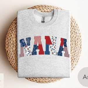 Peut inclure: Un sweat-shirt gris clair avec le mot "NANA" dans un motif patchwork. Les lettres sont rouges, blanches et bleues avec des motifs floraux, cachemire et vichy. Le sweat-shirt est posé sur une surface circulaire tissée.