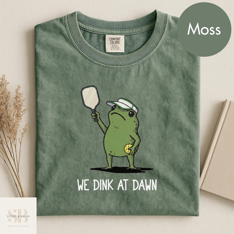 K&ouml;nnte beinhalten: Moosgr&uuml;nes T-Shirt mit einer Cartoon-Frosch, der Pickleball spielt. Der Frosch tr&auml;gt ein Visier und h&auml;lt einen Schl&auml;ger und einen Ball. Der Text "WE DINK AT DAWN" ist auf dem Shirt aufgedruckt. Hergestellt aus Comfort Colors.