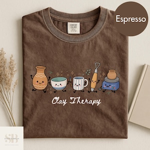 Könnte beinhalten: Ein dunkelbraunes T-Shirt mit Rundhalsausschnitt und einer Grafik aus animierten Töpferwerkzeugen und Gefäßen mit lächelnden Gesichtern. Der Text "Clay Therapy" ist unter den Illustrationen gedruckt. Das Shirt hat ein Comfort Colors Etikett.