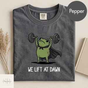 Könnte beinhalten: Ein anthrazitfarbenes T-Shirt mit einer Grafik eines grünen Frosches, der einen schwarzen Umhang trägt und eine Langhantel über den Kopf hebt. Unter dem Frosch steht der Text WE LIFT AT DAWN in weißen Großbuchstaben. Mit Comfort Colors Etikett.