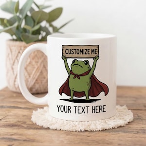Peut inclure: Un mug en céramique blanche illustré d'une grenouille verte de dessin animé portant une cape rouge et tenant un panneau en bois avec l'inscription CUSTOMIZE ME. Sous la grenouille, le texte YOUR TEXT HERE est imprimé en noir.