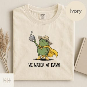 May include: Ivory-colored t-shirt featuring a cartoon frog dressed as a superhero, holding a watering can. The frog wears a hat, cape, and boots. The text "WE WATER AT DAWN" is printed below the frog.