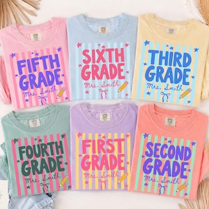 Op de afbeelding: Zes t-shirts in pastelkleuren met verticale strepen en tekst over schooljaren. Elk shirt toont een leerjaar, de naam Mrs. Smith, een potloodillustratie en kleine sterretjes. De shirts zijn roze, lichtblauw, geel, groen, paars en koraalkleurig.