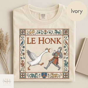 Könnte beinhalten: Ein elfenbeinfarbenes T-Shirt mit einer Grafik im mittelalterlichen Stil und dem Text LE HONK. Die Abbildung zeigt eine Gans mit Krone, die einen Ritter in Kettenrüstung jagt, umrahmt von einer dekorativen Bordüre mit Blumenmotiven und Fabelwesen.