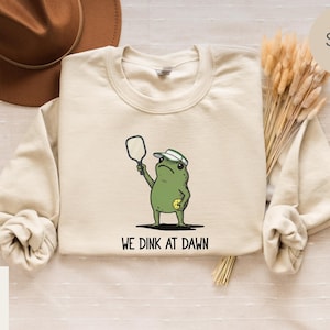 K&ouml;nnte beinhalten: Ein sandfarbenes Sweatshirt mit einem Cartoon-Frosch, der Pickleball spielt. Der gr&uuml;ne Frosch tr&auml;gt einen wei&szlig;en Hut und h&auml;lt einen Schl&auml;ger. Der Text "WE DINK AT DAWN" ist darunter aufgedruckt. Im Hintergrund ist ein brauner Hut zu sehen.