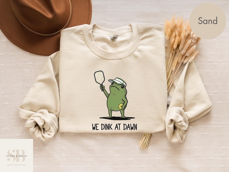 K&ouml;nnte beinhalten: Ein sandfarbenes Sweatshirt mit einem Cartoon-Frosch, der Pickleball spielt. Der gr&uuml;ne Frosch tr&auml;gt einen wei&szlig;en Hut und h&auml;lt einen Schl&auml;ger. Der Text "WE DINK AT DAWN" ist darunter aufgedruckt. Im Hintergrund ist ein brauner Hut zu sehen.