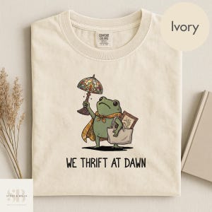 May include: Ivory-colored t-shirt featuring a cartoon frog holding a stained glass lamp and a tote bag. The frog wears a cape and scarf. The text "WE THRIFT AT DAWN" is printed below the frog.