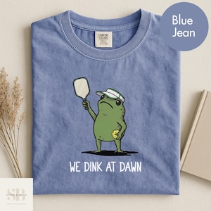 K&ouml;nnte beinhalten: Blaues Jeans-T-Shirt mit einer gr&uuml;nen Froschfigur, die einen Schirmm&uuml;tze tr&auml;gt und einen Pickleball-Schl&auml;ger und -Ball h&auml;lt. Der Text "WE DINK AT DAWN" ist darunter gedruckt. Das Shirt hat ein Comfort Colors-Etikett.