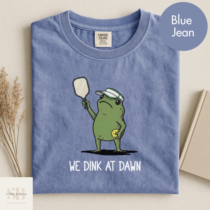 K&ouml;nnte beinhalten: Blaues Jeans-T-Shirt mit einer gr&uuml;nen Froschfigur, die einen Schirmm&uuml;tze tr&auml;gt und einen Pickleball-Schl&auml;ger und -Ball h&auml;lt. Der Text "WE DINK AT DAWN" ist darunter gedruckt. Das Shirt hat ein Comfort Colors-Etikett.