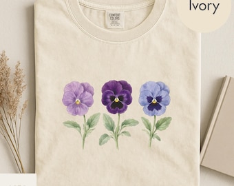 May include: Ivory-colored t-shirt featuring a watercolor design of three pansy flowers in shades of purple and blue. The flowers have green stems and leaves. The shirt has a Comfort Colors tag. The word "Ivory" is in a circle in the upper right corner.