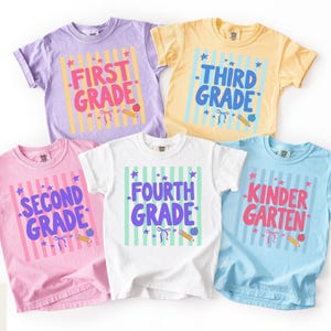 Peut inclure: Cinq t-shirts à manches courtes en lavande, jaune, rose, blanc et bleu clair. Chaque t-shirt présente un fond à rayures verticales avec des graphismes sur le thème de l'école et le texte First Grade, Third Grade, Second Grade, Fourth Grade et Kindergarten.