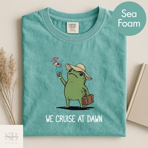 May include: Seafoam green t-shirt featuring a cartoon frog wearing a hat and holding a cocktail and suitcase. The text "WE CRUISE AT DAWN" is printed below the frog. The shirt has a crew neck and a Comfort Colors tag.