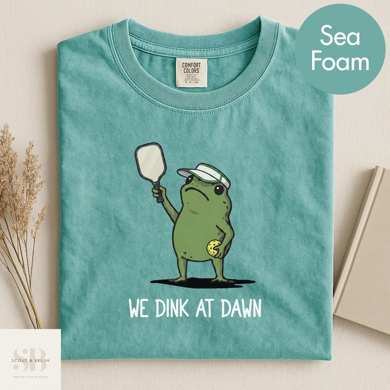 K&ouml;nnte beinhalten: Meergr&uuml;nes T-Shirt mit einem Cartoon-Frosch, der Pickleball spielt. Der Frosch tr&auml;gt ein Visier und h&auml;lt einen Schl&auml;ger und einen gelben Ball. Der Text "WE DINK AT DAWN" ist darunter gedruckt.