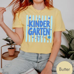 May include: A butter yellow short-sleeve t-shirt featuring a graphic print with light blue vertical stripes, blue stars, a blue bow, a yellow pencil, and a blue apple. The text "KINDER GARTEN" is printed in large, bold blue letters across the center.