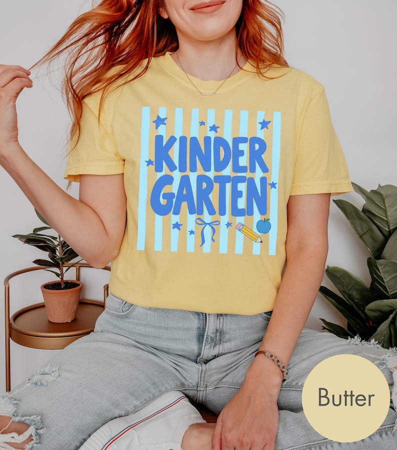 May include: A butter yellow short-sleeve t-shirt featuring a graphic print with light blue vertical stripes, blue stars, a blue bow, a yellow pencil, and a blue apple. The text "KINDER GARTEN" is printed in large, bold blue letters across the center.