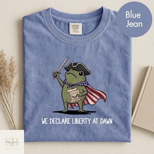 Könnte beinhalten: Blaues Jeans-T-Shirt mit einer Cartoon-Froschfigur, die einen Dreispitzhut trägt, ein Schwert und eine Schriftrolle hält. Der Frosch trägt einen Umhang mit roten und weißen Streifen und einen blauen Schal mit weißen Sternen. Der Text "WE DECLARE LIBERTY AT DAWN" ist darunter gedruckt.