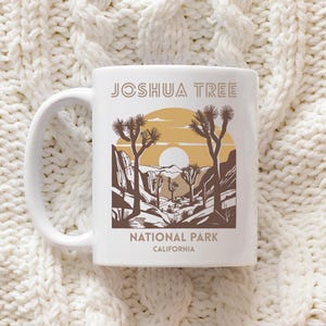 May include: White ceramic mug featuring a desert landscape design with the text "Joshua Tree National Park California." The design includes a sun, Joshua trees, and mountains in shades of brown and tan.