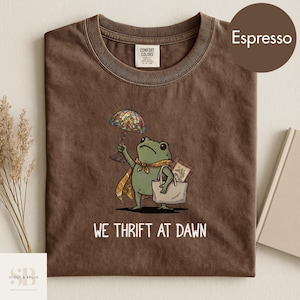 Może przedstawiać: T-shirt w kolorze espresso z rysunkiem żaby trzymającej lampę witrażową i torbę na zakupy. Pod spodem wydrukowano napis "WE THRIFT AT DAWN". Koszulka wykonana jest z miękkiego materiału.