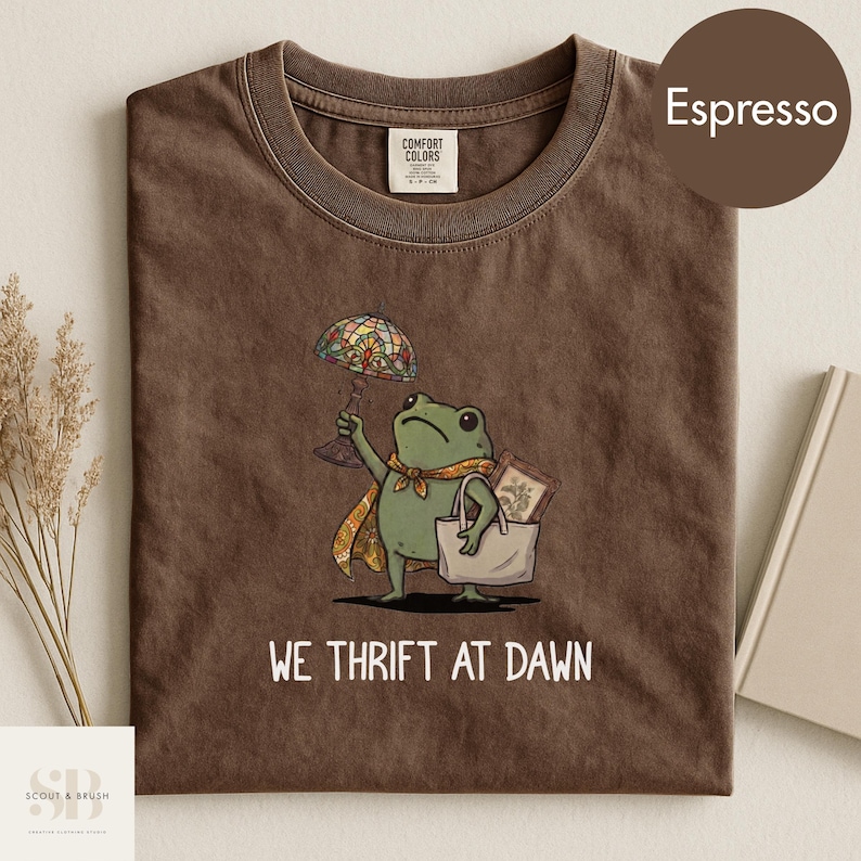 Może przedstawiać: T-shirt w kolorze espresso z rysunkiem żaby trzymającej lampę witrażową i torbę na zakupy. Pod spodem wydrukowano napis "WE THRIFT AT DAWN". Koszulka wykonana jest z miękkiego materiału.