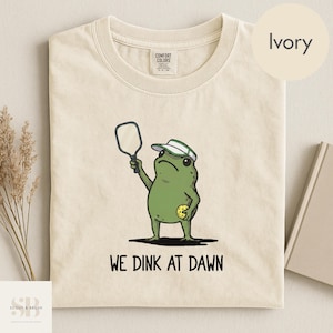 K&ouml;nnte beinhalten: Elfenbeinfarbenes T-Shirt mit einer Cartoon-Froschfigur, die einen Schirmm&uuml;tze tr&auml;gt und einen Pickleball-Schl&auml;ger und -Ball h&auml;lt. Der Text "WE DINK AT DAWN" ist darunter gedruckt. Das Shirt ist aus weichem Material.