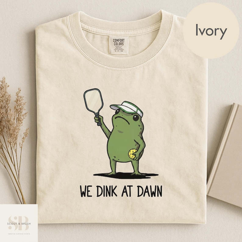 K&ouml;nnte beinhalten: Elfenbeinfarbenes T-Shirt mit einer Cartoon-Froschfigur, die einen Schirmm&uuml;tze tr&auml;gt und einen Pickleball-Schl&auml;ger und -Ball h&auml;lt. Der Text "WE DINK AT DAWN" ist darunter gedruckt. Das Shirt ist aus weichem Material.
